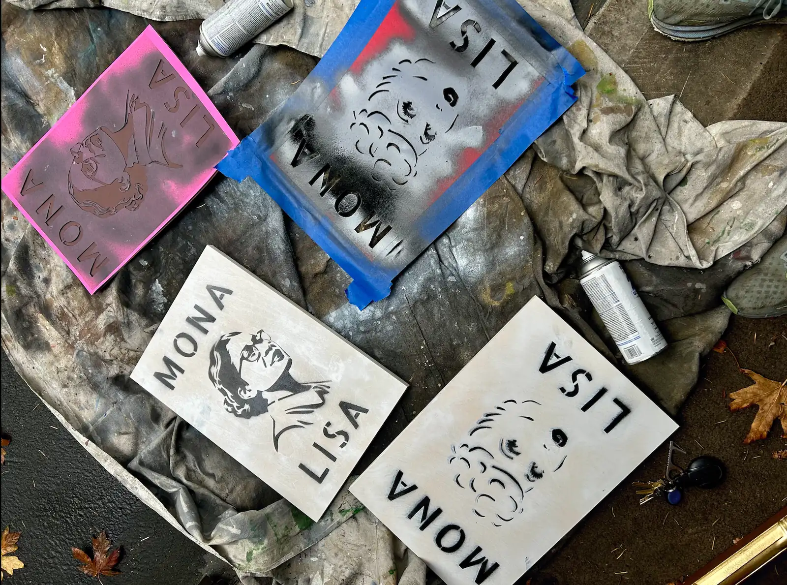 Freshly sprayed MONA LISA boards and their cut stencils on a paint-stained drop cloth, with spray cans, blue tape and fallen leaves